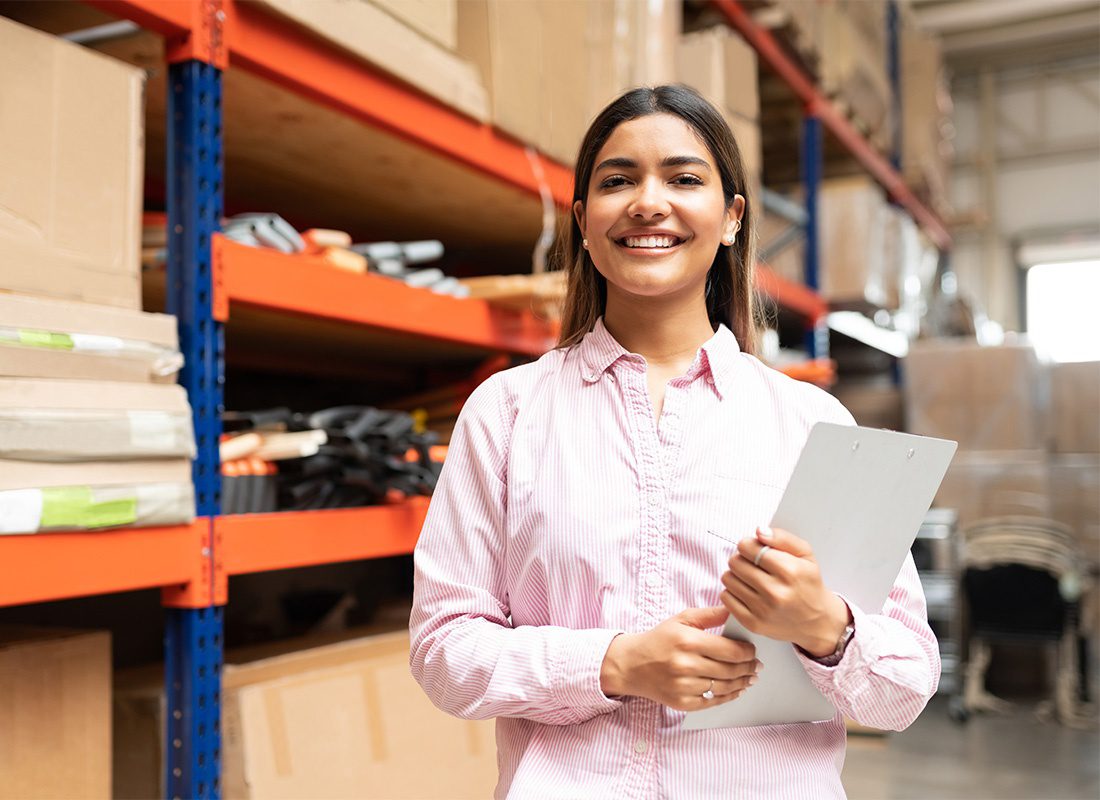 Business Insurance - Smiling Portrait of a Young Female Warehouse Manager in a Button Down Shirt Holding Paperwork in her Hands While Standing Next to Large Shelves Full of Inventory